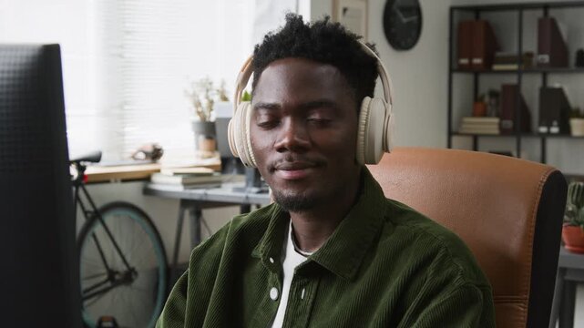 Medium close-up portrait of focused gen Z African American male web developer, wearing wireless headphones, working on project code on desktop computer in office, looking at camera, smiling
