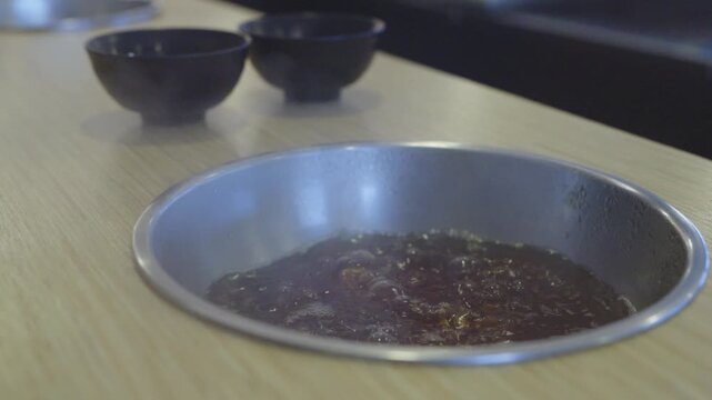 Boiling dark broth inside a metal pot recessed into a wooden table at a restaurant. Two empty black bowls sit in the background. Traditional Asian hot pot or shabu-shabu dining experience.