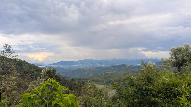 A vast, tranquil landscape overlooking dense forests and rolling mountain ridges in the Sri Lankan highlands. Low-lying mist and a distant glowing horizon highlight the natural beauty under a moody, o