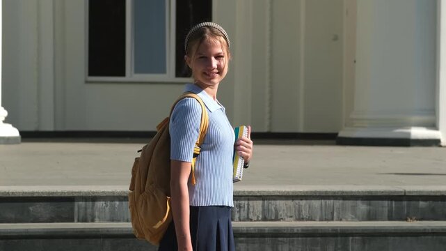 Portrait of smiling teenage girl in uniform with backpack holding books outdoors on building background. Concept of happy student and back to school