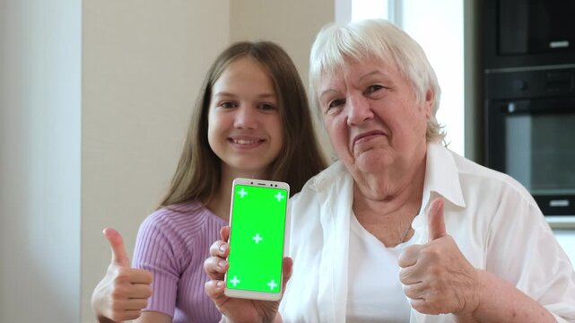 Granddaughter and grandmother holding smartphone with vertical green screen and showing thumbs up. Concept of mobile application and user satisfaction.