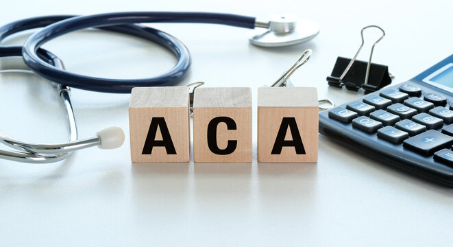 Wooden blocks spell ACA. Stethoscope rests beside them. Calculator and clips sit nearby. Office desk surface is clean and white.