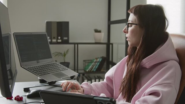 Medium side shot of young Caucasian female developer, wearing cozy pink sweatshirt, typing algorithm on desktop computer, testing on laptop and tablet while working on mobile app with vibe coding