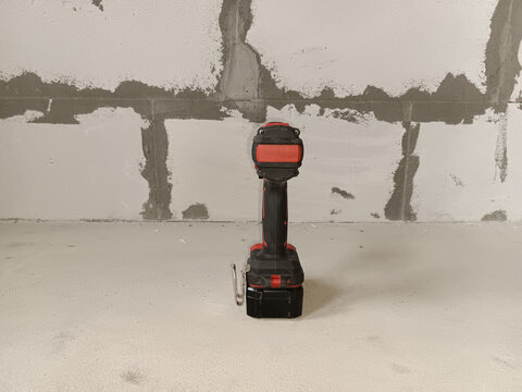 A black and red cordless drill rests on a concrete surface. The background features unfinished drywall with visible seams and patches. Back view