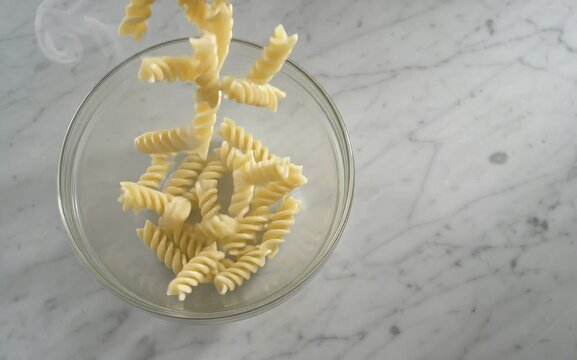 Steaming fusilli pasta dropping into a glass bowl on marble table