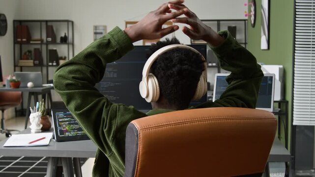 Medium close-up of professional black male programmer sitting back in chair with hands clasped on back of head, relaxing while watching vibe coding agent writing script for current mobile app project