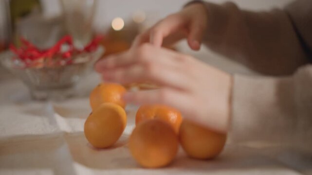 Closeup hands arranging mandarins on table, woman in knit sweater organizes fruit into line, soft bokeh lights, warm color palette, tactile slow motion, gentle fidgeting, holiday centerpiece