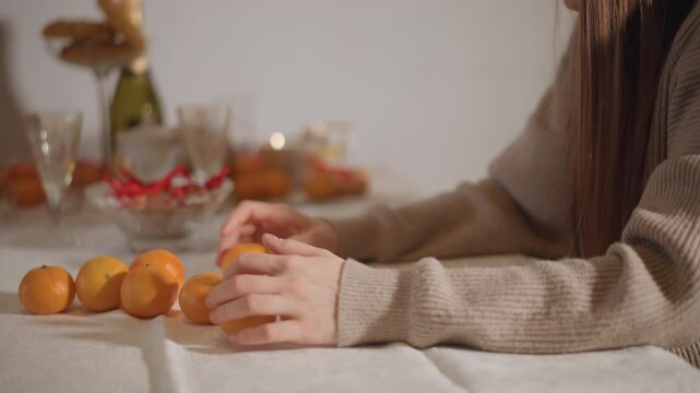 Side view woman lining mandarins on table, longhaired in knit sweater arranges fruit in row beside glassware and wrapped gifts, deliberate motion, domestic holiday prep, warm mood