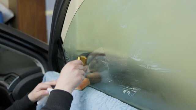 Automotive technician carefully cleaning and preparing a car side window with a sponge and soapy water before applying a protective tint film in a professional auto detailing service garage
