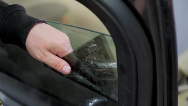 Close-up shot of a skilled male worker carefully removing old, bubbled tinting film from a car's side window using steam from a heat gun, preparing the glass for a new application