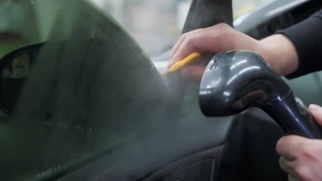 Professional detailer carefully removing tint film to a car window, using a yellow squeegee and a steamer for a smooth, bubble-free installation in a specialized automotive workshop