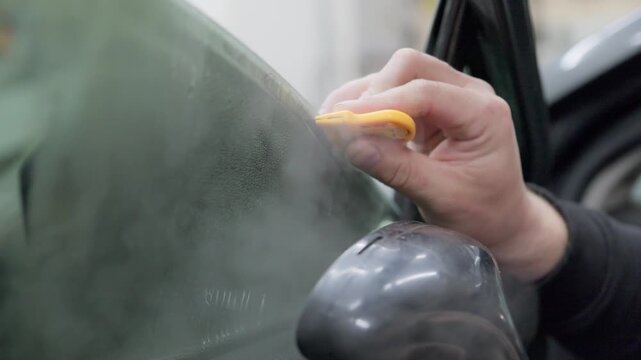 Professional auto service worker carefully removing old tinting film from a car's side window using a hot steam generator and a plastic scraper, preparing the glass for new tint installation