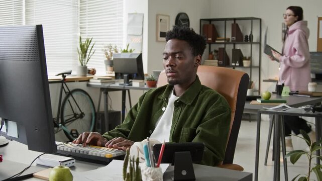 Medium shot of African American male software developer working on computer, junior Caucasian female colleague approaching with laptop to ask advice on code, while collaborating on project in office