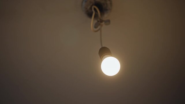 Bright hanging bulb over aged socket, soft yellow glow illuminates textured ceiling, twisted cord loop, subtle hum, maintenance vibe, electrician action implied, candid household detail