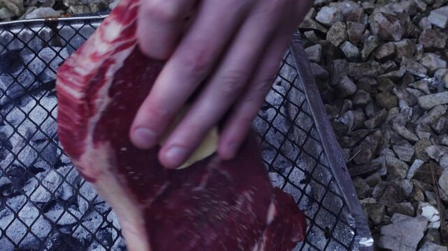 Close up of hand placing a raw beef steak with a knob of butter onto a disposable charcoal grill grate outdoors