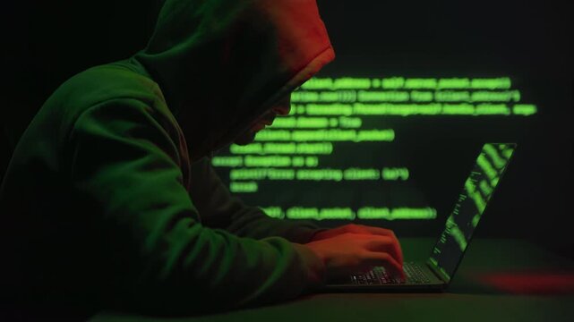 Hooded anonymous hacker typing on laptop in dark room with green code on screen and background. Cyber attack, data breach, malware activity and online security threat concept.