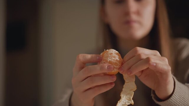 Caucasian woman peeling mandarin with focus closeup on hands removing rind delicate segments exposed juice glistening neutral kitchen background evening snack ritual mindful eating tactile detail slow