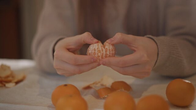 Hands holding mandarin halves over table, paired segments separated gently with surrounding whole mandarins and peels, sweater cuff visible, warm soft lighting, offering gesture and intimate home