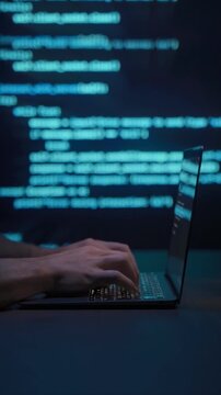 Close up of programmer hands typing on laptop keyboard in dark workspace with blue code lines in background. Coding process and software development or cybersecurity concept. Vertical video