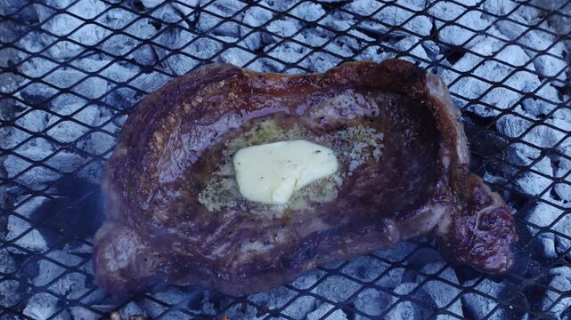 Close-up of a cooked beef steak with a melting pat of butter on top resting on a charcoal grill grate outdoors