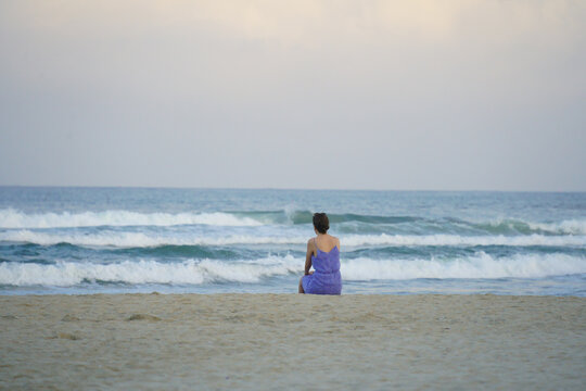 Rear view of a serene woman in a purple sundress sitting on a sandy beach and looking at ocean waves during overcast day