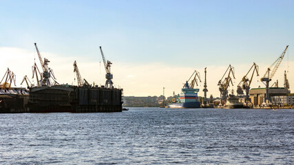 Obraz premium Saint Petersburg, Russia: Wide view from Blagoveshchensky Bridge to Baltic Shipyard with cranes, dockyards and cargo vessel on Neva River. Industrial shipping and logistics scene