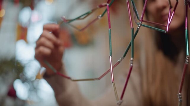 Hands crafting metallic holiday garland with colorful tubes, closeup of fingers threading beads, blurred tree background, warm natural light, sweater sleeve, gentle motion, tactile focus, festive