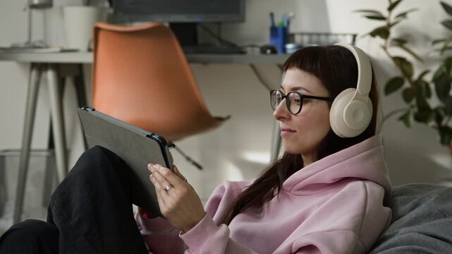 Medium close-up of young Caucasian female developer reviewing script provided by vibe coding agent, using tablet computer and stylus, while enjoying upbeat music in headphones, relaxing in bean bag