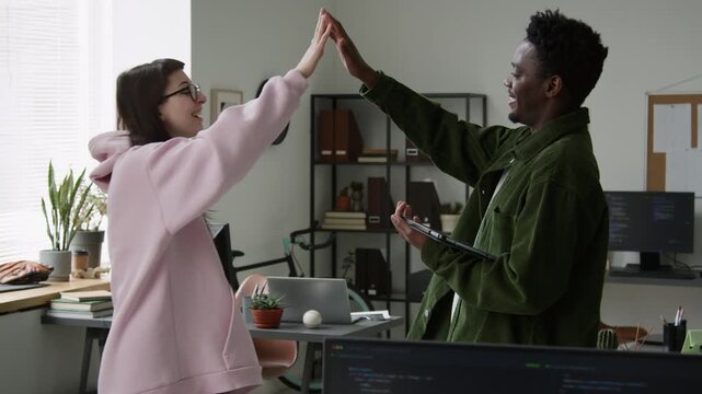 Medium shot of Caucasian female and African American male software engineers discussing plan for work on mobile app project, smiling and giving high fives at successful collaboration