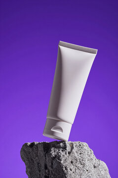 Cosmetic tube stands tilted on a rugged gray stone base against a solid violet backdrop. Harsh side light emphasizes the smooth plastic surface and coarse mineral texture.