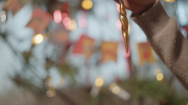 Gold icicle ornament held against bokeh soft sweater sleeve, colorful garland and warm string lights, gentle placement into evergreen branch, cozy nostalgic atmosphere, slow cinematic movement,