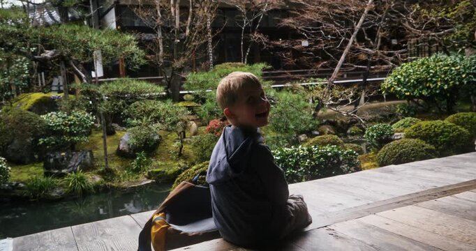 Little boy with backpack sitting on old Japanese home deck near garden then standing up - steady cam shot