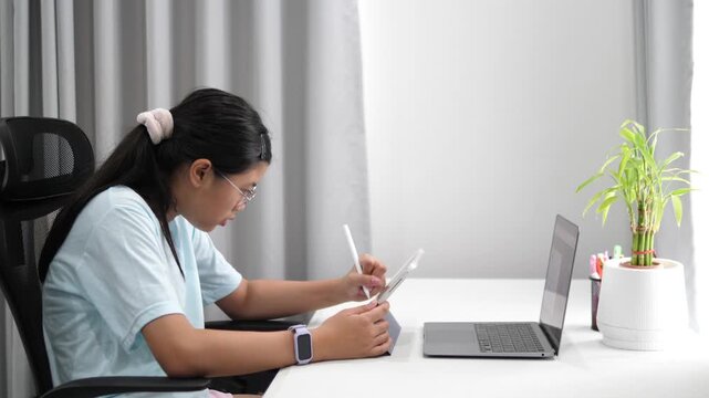 Cinematic side profile shot of a young girl with glasses studying online, using a stylus pen on tablet with a laptop on a white desk