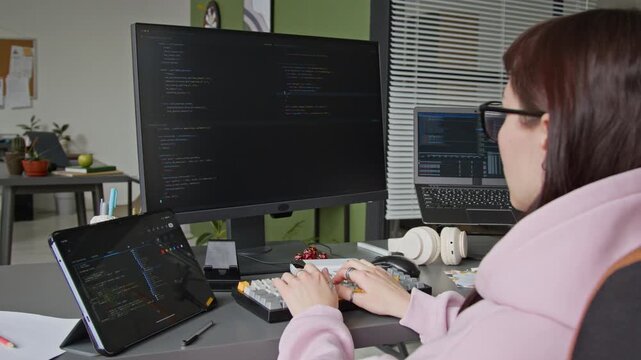 Medium over-shoulder of young female software developer writing algorithm on computer with vibe coding assistant, laptop and tablet on desk, while male colleague throwing darts in background