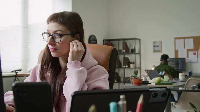 Medium shot of cheerful Caucasian female software developer putting in earphones, turning on favorite music on smartphone for inspiration then continuing to work on code while swaying to rhythm