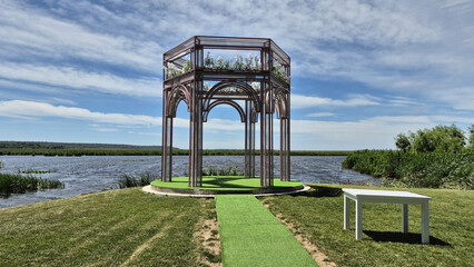 An elegant, modern metal gazebo stands decorated with white flowers on a lakeside grassy lawn under a partly cloudy sky, prepared for an outdoor event. © aura