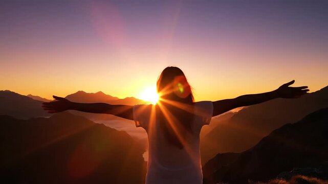 Woman stands atop a mountain peak with arms outstretched, embracing the sunrise and feeling a profound sense of freedom and inner peace.