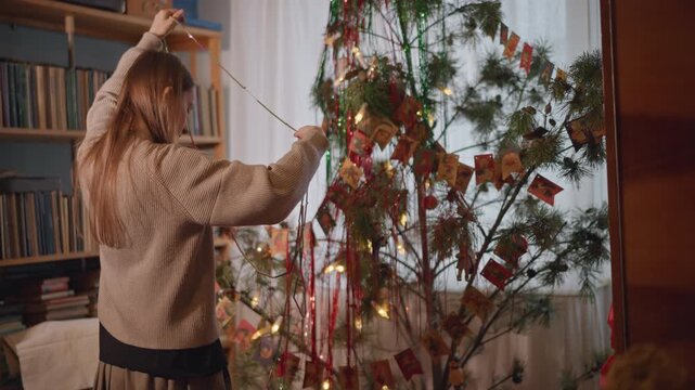 White woman strings lights on studio tree by bookshelf and records, arranging handmade ribbons and paper cards indie musician ritual in warm evening light preparing seasonal display with creative