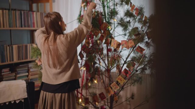 White woman decorates cozy living room tree with handmade garlands and warm lights, beside bookshelf and vinyl collection intimate vintage holiday scene as family archivist arranges sentimental