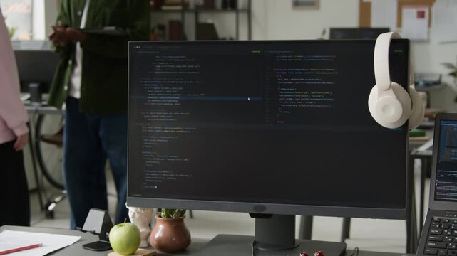 Medium shot of vibe coding assistant working on computer, writing script, while two unrecognizable web developers discussing project implementation plan in background, demonstrating automation