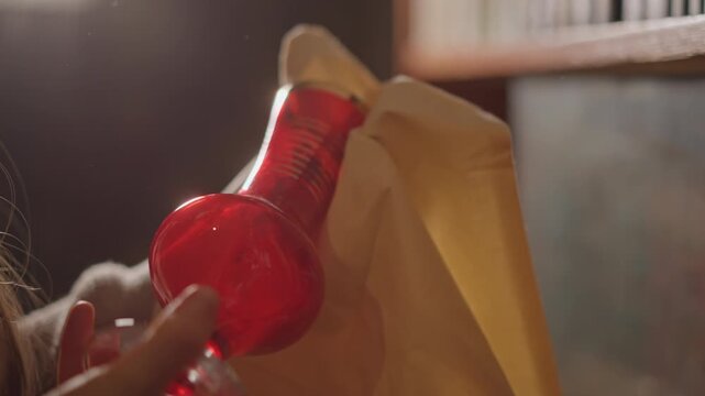 Caucasian woman unboxing red glass vase from parcel in cozy library, hands gently reveal fragile vintage piece under warm golden light, intimate closeup showing careful handling and provenance hints