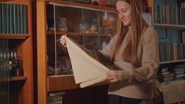 Caucasian woman unfolding heirloom cloth. Cozy study scene with wooden cabinet, bookshelf backdrop, gentle sweater, careful hands revealing porcelain and glass collectibles, warm golden light,