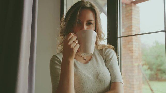 Blonde hair young happy woman holding cup of hot natural tea, coffee in hands drinking enjoys aroma of drink. Beauty smiling face adult girl cozy room home near window full fog, cozy casual wear 