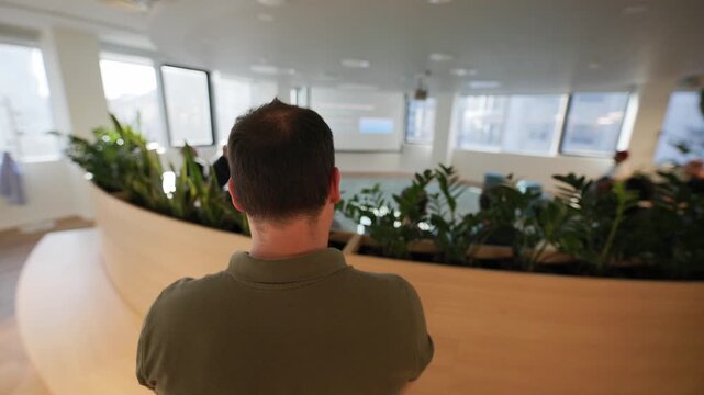 Male participant in a modern office setting observes a presentation, surrounded by indoor plants and colleagues seated in a collaborative workspace
