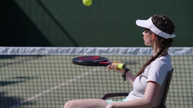 Young woman with physical disability sitting in a chair and playing padel tennis. Slow motion view of her hitting the ball with the racket