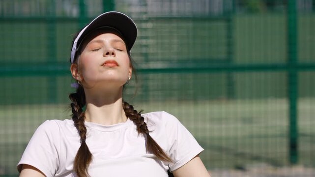 Portrait of a young female athlete in a visor sitting on a tennis court. Tired sportswoman resting and recovering after the game