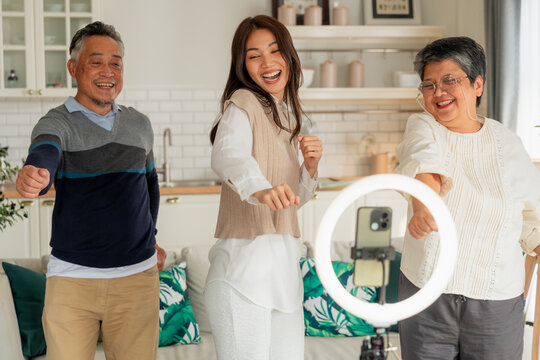 Happy Asian multi-generation family making vlog video live streaming online with smart phone and ring light inside home kitchen concept for social media influencer digital content creator technology m