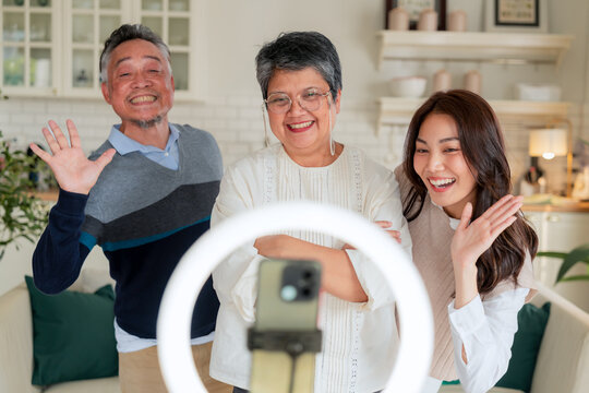 Happy Asian multi-generation family making vlog video live streaming online with smart phone and ring light inside home kitchen concept for social media influencer digital content creator technology m