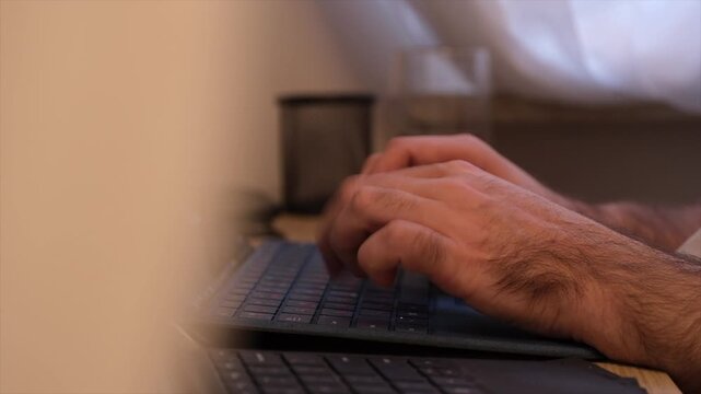 Close up view of a man typing on a laptop while working from home. Warm indoor atmosphere with a cup of tea in the background creates a realistic remote work environment. Ideal for concepts of freelan