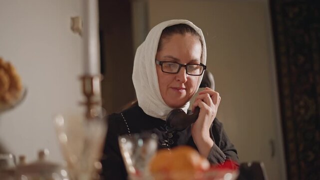 White woman in headscarf at dining table speaking on telephone, warm candlelit setting with crystal glassware and fruit centerpiece, attentive expression, hostess role, family conversation, cozy